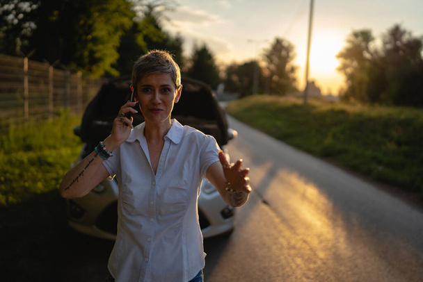 one woman mature female standing on the road in the evening sunset by the broken vehicle car automobile failed engine open hood making a phone call for help roadside assistance towing service concept - Foto, afbeelding