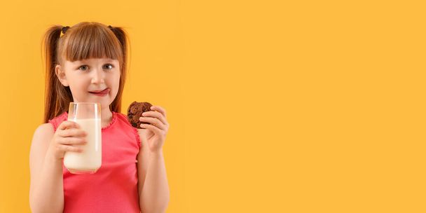 Cute little girl drinking milk and eating tasty cookie on yellow background with space for text - Foto, imagen