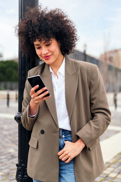 portrait of a smiling young latin woman checking her phone leaning against a city lamppost, technology and communication concept - Фото, зображення