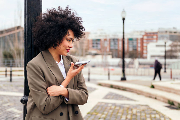smiling young latin woman recording a voice message with her phone leaning against a city lamppost, technology and communication concept, copy space for text - Foto, afbeelding