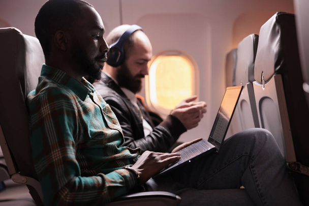 Joven freelancer viajando en viaje de trabajo en avión en clase económica, trabajando en laptop durante el vuelo al atardecer. Empleado masculino volando con vuelo comercial con aerolínea internacional. - Foto, imagen