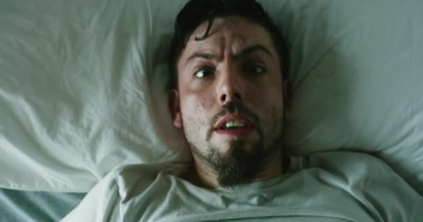 A man suffering from insomnia has a nightmare in bed at night. A scared man gets frighted and looks terrified after having a bad, horrific dream. Guy wakes up and panics while lying in a dark room. - Video, Çekim