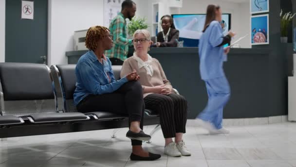 Diverse female patients sitting in hospital waiting area, talking about healthcare to attend medical appointment. People waiting in facility reception to start consultation with disease specialist. - Footage, Video