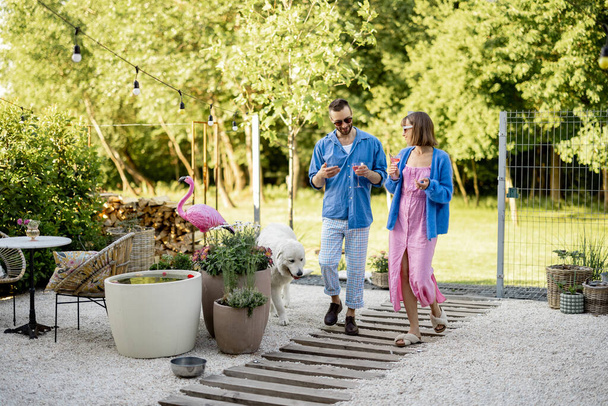 Young stylish couple have romantic conversation while walking with a drink at their beautiful backyard of country house. Young man and woman spend summer time happily - Fotografie, Obrázek