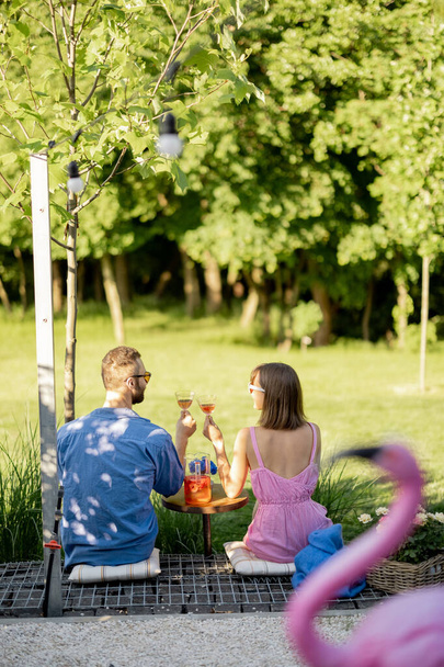 Young couple sit on porch of country house, have a drink and enjoy beautiful view on nature, view from the backside. Man and woman spend summer time romantically - Photo, Image