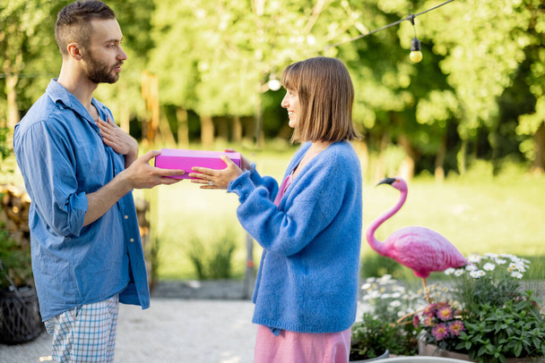 Man geeft een geschenk in roze doos aan zijn vrouw, vieren verjaardag in de tuin van hun huis. Viering van dankdag, Valentijnsdag of verjaardagsconcept - Foto, afbeelding