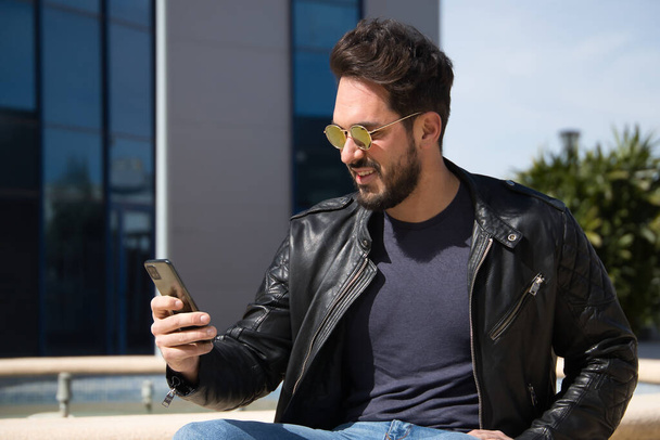 Handsome young man with beard, sunglasses, leather jacket and jeans, sitting, checking social networks on his cell phone. Concept beauty, fashion, trend, app, smartphone, social networks. - Photo, Image