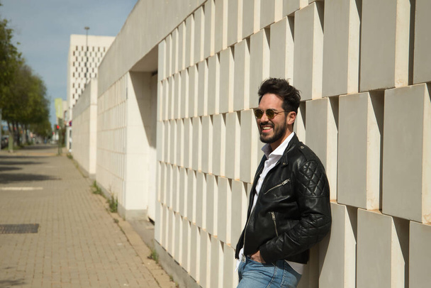 Handsome young man with beard, sunglasses, leather jacket, white shirt and jeans, leaning against a wall smiling very happy. Concept beauty, fashion, trend, happiness, laughter. - Fotoğraf, Görsel
