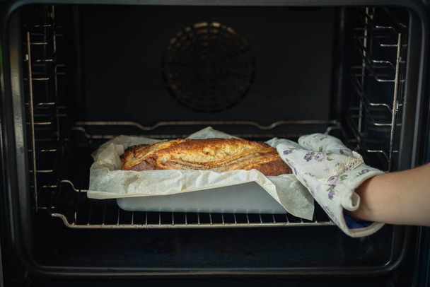 Woman wearing potholder taking fresh banana bread out of the oven. Close up of hot homemade pastry - Foto, Imagem