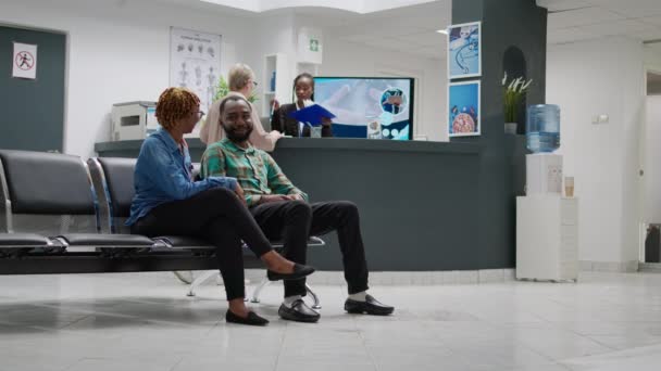 Homme et femme afro-américain parlant de maladie dans le hall d'accueil de l'hôpital, attendant d'assister à un rendez-vous de consultation avec un spécialiste. Les patients dans la salle d'attente. Trépied. - Séquence, vidéo
