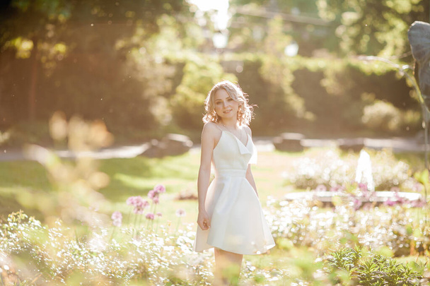 Joven hermosa rubia de 20 años con el pelo rizado en un vestido blanco en los soleados rayos del atardecer en el parque. Un modelo elegante camina por el parque floreciente de las ciudades en verano. Verano. - Foto, Imagen