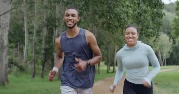 Two fit people running in a park, enjoying their daily exercise and fitness routine outdoors. Confident smiling female jogging and happy african male working out in a scenic forest in nature. - Felvétel, videó