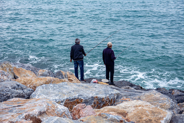 Balıkçılar bulutlu bir günde deniz kenarındaki taşların üzerinde otururken balık tutuyorlar. Denizde ve gökyüzünde ağır bulutlarla küçük dalgalar. - Fotoğraf, Görsel