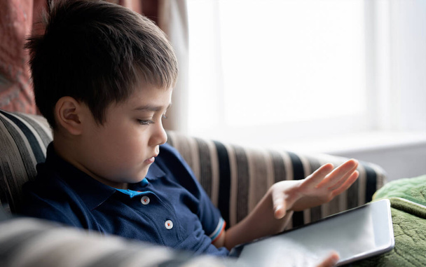 Cinematic portrait young boy playing game on tablet sitting on sofa with light shining from window, Kid playing games online on internet at home, Child talking video call with friends at home - Foto, imagen