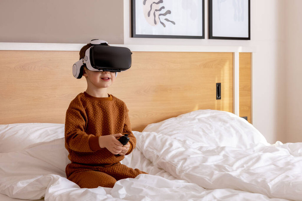 Little adorable boy sitting on bed at home with VR headset and playing interactive video game, exploring virtual reality. Cute child wearing VR glasses. Future, gadgets, technology, education online - Фото, зображення