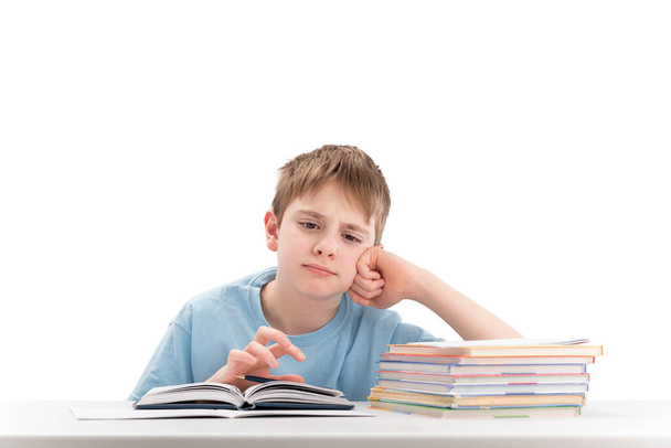 Tired boy doing homework at table and attentively reads book. Portrait of schoolboy at desk with stack of notebooks. White background - Zdjęcie, obraz