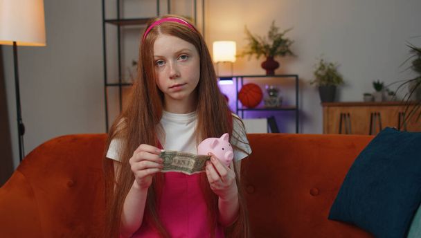 Poor young teen child girl insufficient amount of money, holding piggybank and one dollar banknote at home. Financial crisis. Bankruptcy. Poverty and destitution. Ginger kid in living room on couch - Fotoğraf, Görsel