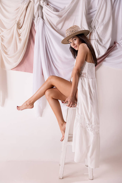 Portrait of stylish woman sitting on chair, posing in white dress and straw hat isolated over textile background. Concept of natural beauty, fashion, wellness, lifestyle, femininity, summer style, ad - Foto, Bild