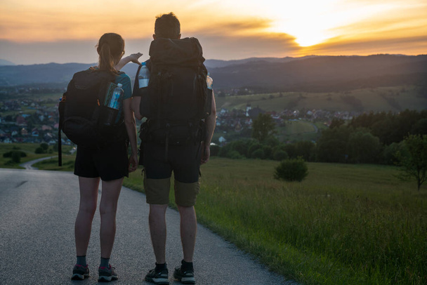 A pair of tourists hiking along a beautiful scenic road in the mountains enjoying the sunset - Zdjęcie, obraz