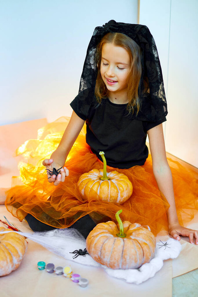 Ребенок играет с пауком, украшает тыкву дома, маленькая девочка рисует лицо на оранжевой тыкве Хэллоуина. Концепция праздничного оформления - Фото, изображение