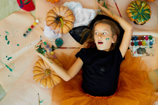 Vue du dessus de Souriante fille couchée sur le sol entre dessiné citrouille Halloween, et la décoration pour la fête, les mains de peinture, l'émotion, faire des visages. - Photo, image