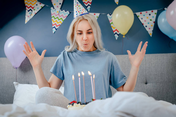 Happy woman in pajama and party cap blowing out candles on birthday cake on the bed in decorated bedroom. Make a wish process. Morning surprise. Happy birthday concept. Selective focus - Zdjęcie, obraz