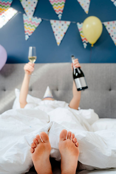 Selective focus on foots of woman lying on bed with raised hands holding bottle and glass of champagne in decorated bedroom. Plans for birthday. Time for yourself. Celebration at home. Happy birthday. - Fotoğraf, Görsel