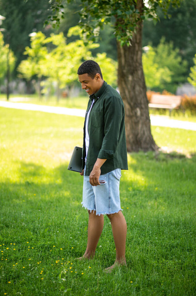 Иди босиком. Темнокожий парень боком к камере идет босиком по траве с ноутбуком глядя вниз улыбаясь в парке - Фото, изображение
