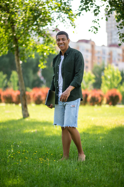 Pleasure. Young dark-skinned man with laptop standing barefoot on grass smiling at camera in park on summer day - Valokuva, kuva