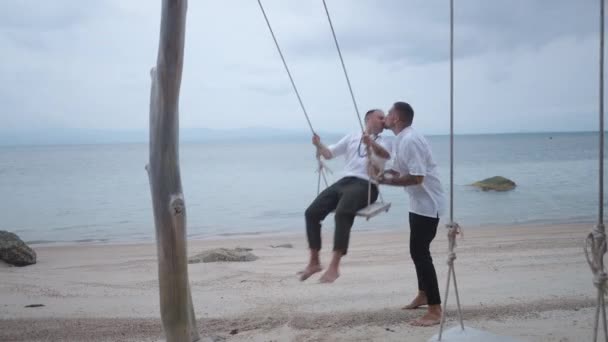 Lovely male gay couple honeymooners kissing while riding a beach swing at tropical island at dusk - 4K Horizontal video - Séquence, vidéo