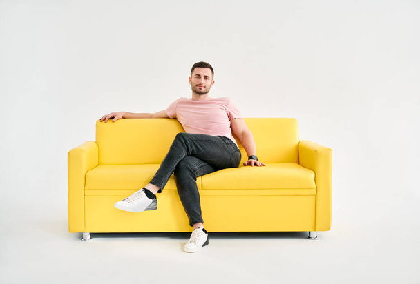 Handsome trendy man posing looking to camera while sitting on comfort sofa over white background. Male portrait - Foto, afbeelding