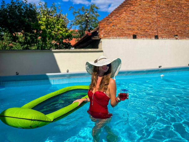 Fiatal lány piros bikiniben napszemüvegben és nyári kalapban szórakozik a medencében, miközben egy italt tart a kezében. - Fotó, kép