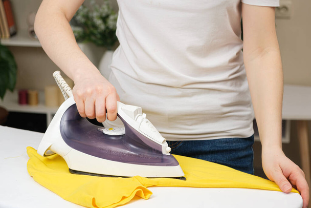 Uma dona de casa passa uma T-shirt amarela com um ferro. Roupa de engomar em casa, cuidar de roupas. Mãos à vista. - Foto, Imagem