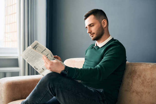 Confident young man reading newspaper and latest news while sitting on sofa in the morning in modern interior - Valokuva, kuva