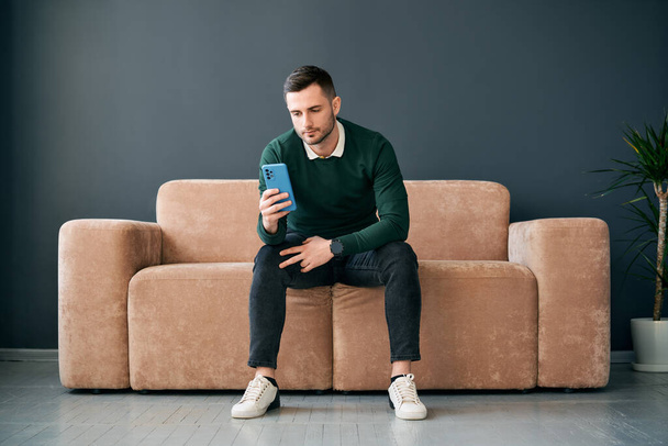 Relaxed man sitting on couch using hsi mobile phone. Social media concept - Фото, изображение