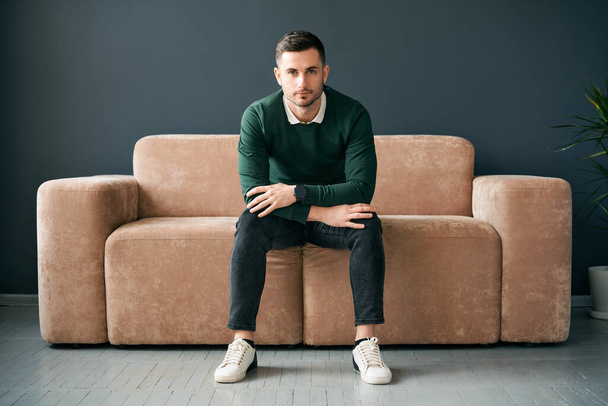 Handsome trendy man posing looking to camera while sitting on comfort sofa in modern apartment. Male portrait - Fotografie, Obrázek