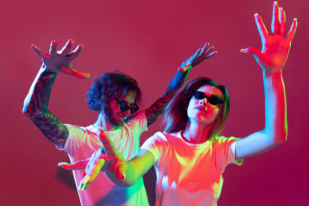 Portret młodego stylowego mężczyzny i kobiety tańczących, pozujących na czerwonym tle w neonowym świetle. Czas na imprezę motywacyjną. Pojęcie młodości, stylu życia, związku, emocji, wyrazu twarzy, reklamy - Zdjęcie, obraz