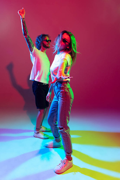 Portrait of young stylish man and woman dancing, posing isolated over red background in neon light. Dancing. Feeling good. Concept of youth, lifestyle, relationship, emotions, facial expression, ad - Photo, Image