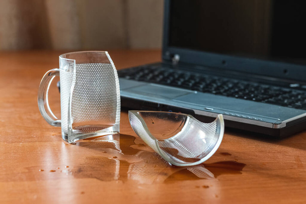 Broken glass mug with spilled coffee or tea near laptop in the office - Fotografie, Obrázek