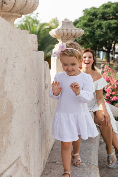Улыбающийся ребенок идет по каменной скамейке моста Пуэнте-дель-Мар рядом с размытой мамой в Валенсии - Фото, изображение