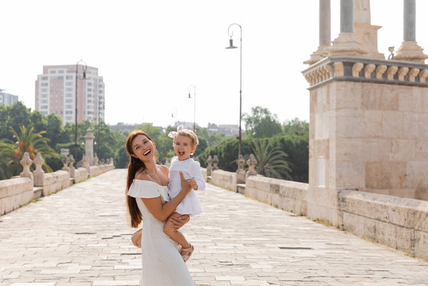 Happy woman holding baby in white dress and looking at camera on Puente Del Mar bridge in Valencia - Foto, immagini