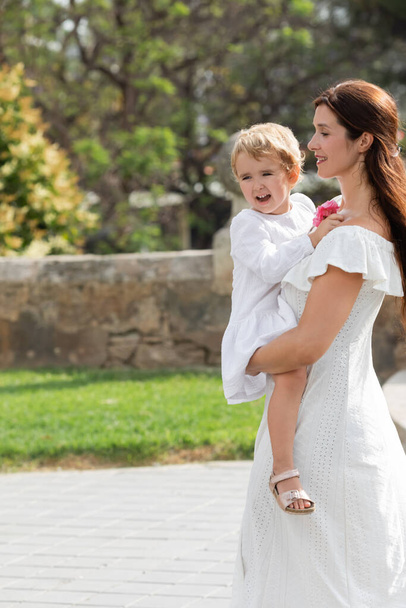 Woman in white dress carrying toddler daughter with flowers in Valencia - Photo, Image
