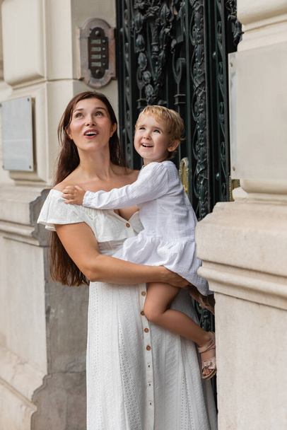 Excited mother and daughter in summer dresses looking away near building in Valencia - Foto, Imagem