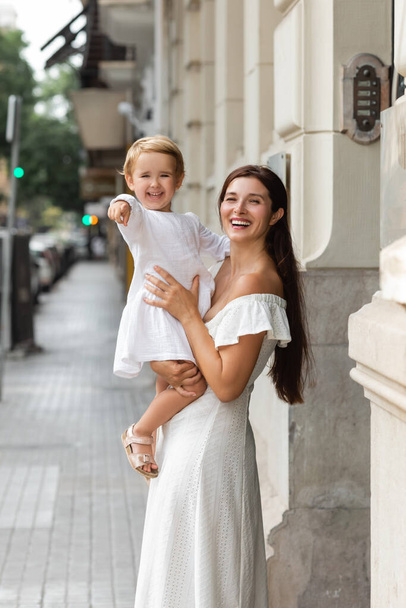 Cheerful brunette woman holding daughter pointing with finger on urban street in Valencia - Photo, image