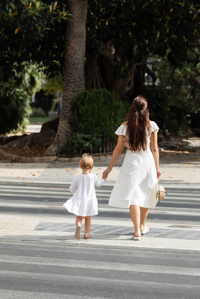 Rückansicht einer Frau mit Sonnenhut, die mit ihrer kleinen Tochter auf einem Fußgängerüberweg in Valencia spazieren geht - Foto, Bild
