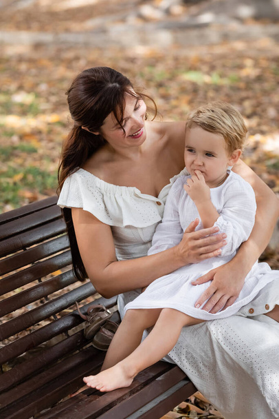Smiling woman in dress hugging toddler daughter on bench in park  - Photo, image