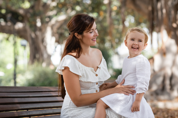 Brunette woman looking at daughter laughing on bench in park - Foto, imagen