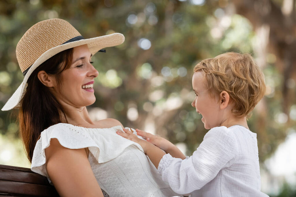 Toddler girl touching smiling mom in sun hat in park  - Photo, image