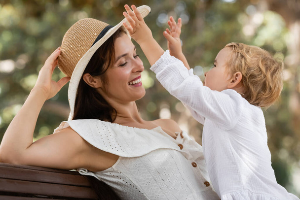Niña jugando con mamá en el sombrero de verano en el banco en el parque  - Foto, Imagen