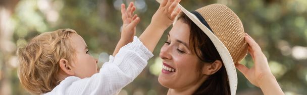 Toddler child touching straw hat on smiling mom in summer park, banner  - Fotó, kép